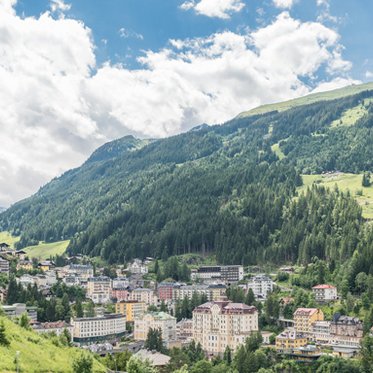 Panoramaaufnahme von Bad Gastein. Die Häuser liegen mitten in den Hängen, es ist ein schöner Sommertag, die Hänge sind grün, Wolken am blauen Himmel.