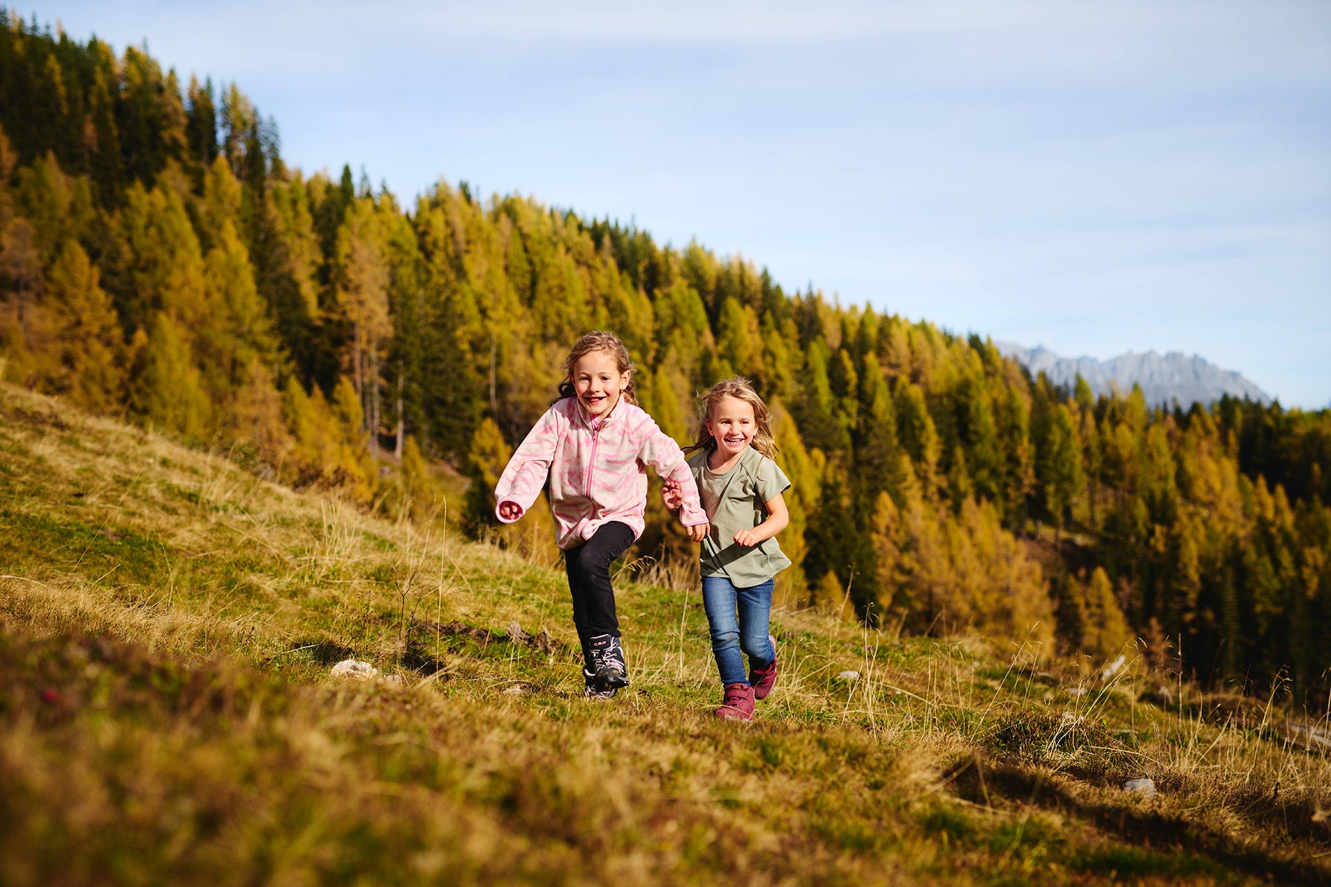 Zwei Kinder laufen lachend über eine herbstliche Almwiese im Gasteinertal, im Hintergrund leuchten goldgelbe Lärchen.