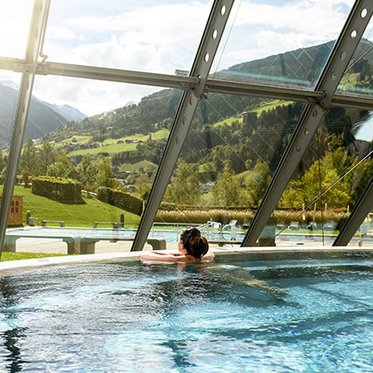 Frau entspannt im Innenbecken der Alpentherme Gastein und blickt durch ein großes Panoramafenster auf die herbstliche Landschaft.