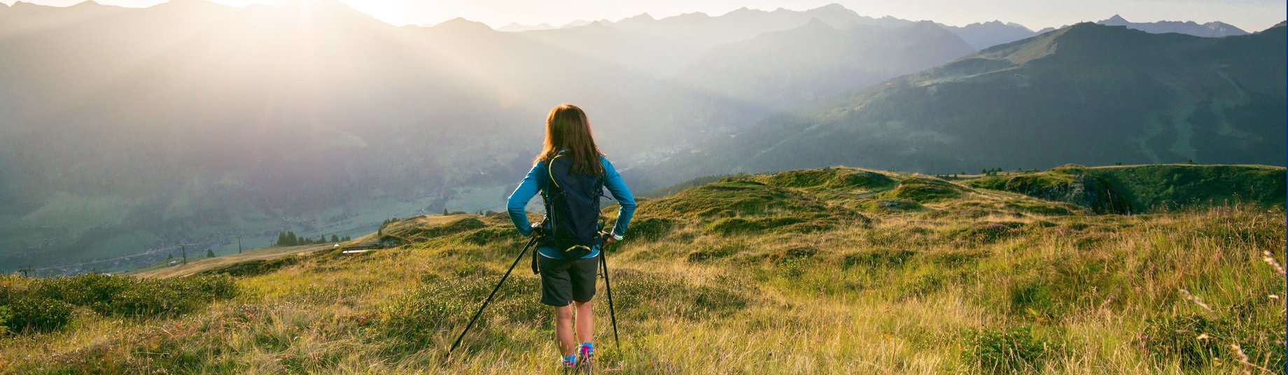 Eine Person mit Rucksack wandert über eine blühende Almwiese im warmen Abendlicht, im Hintergrund erstrecken sich sanfte Bergketten unter einem leicht bewölkten Himmel.