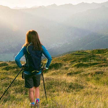 Eine Person mit Rucksack wandert über eine blühende Almwiese im warmen Abendlicht, im Hintergrund erstrecken sich sanfte Bergketten unter einem leicht bewölkten Himmel.