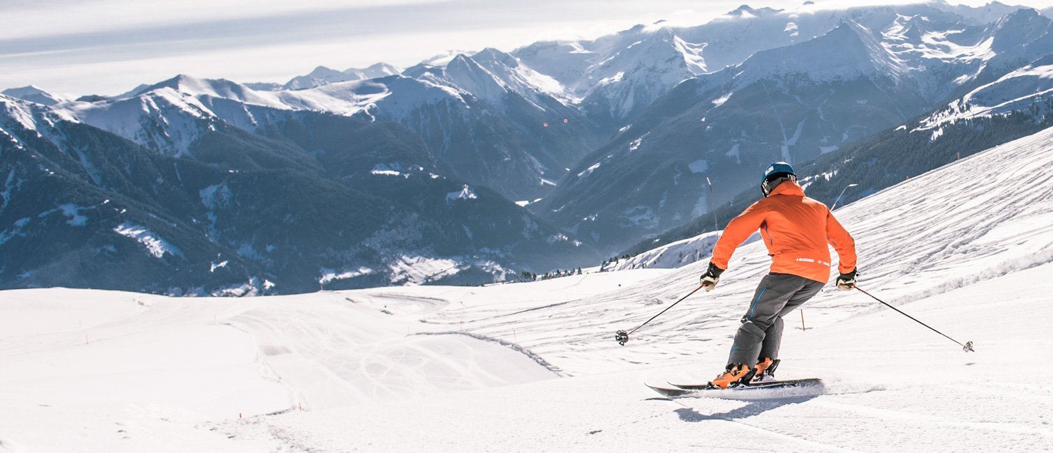 Eine Person in orangefarbener Jacke fährt auf Skiern eine verschneite Bergpiste hinunter, im Hintergrund sind schneebedeckte Gipfel und ein weites alpines Panorama zu sehen.