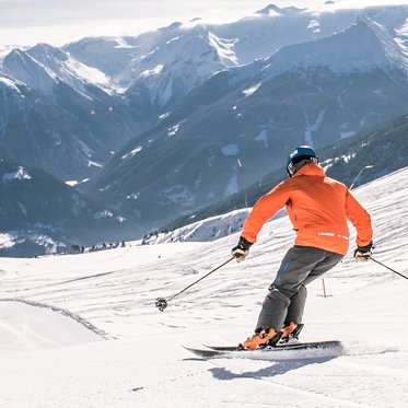 Eine Person in orangefarbener Jacke fährt auf Skiern eine verschneite Bergpiste hinunter, im Hintergrund sind schneebedeckte Gipfel und ein weites alpines Panorama zu sehen.