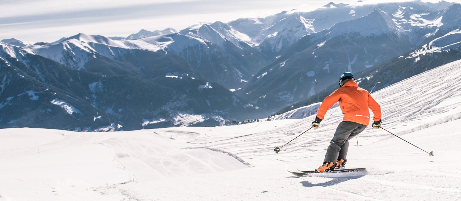 Eine Person in orangefarbener Jacke fährt auf Skiern eine verschneite Bergpiste hinunter, im Hintergrund sind schneebedeckte Gipfel und ein weites alpines Panorama zu sehen.