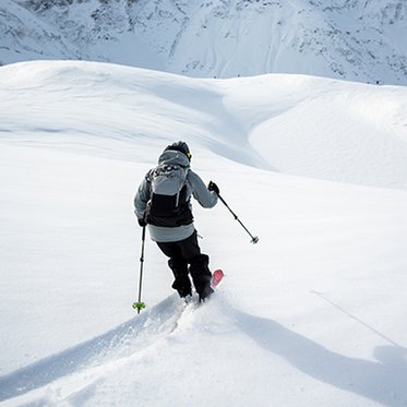 Eine Person mit Rucksack fährt auf Skiern durch unberührten Tiefschnee in einem weiten, verschneiten Hochgebirge; vor ihr breiten sich sanfte Schneehänge unter klarem Himmel aus.