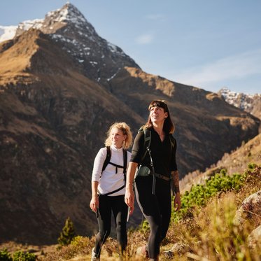 Zwei Frauen mit Rucksäcken wandern auf einem Bergpfad durch eine herbstliche alpine Landschaft mit goldenen Gräsern und Blick auf schroffe, teilweise schneebedeckte Berggipfel.