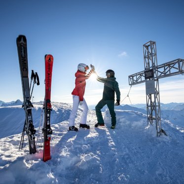 Zwei Personen stehen mit Skiern auf einem verschneiten Berggipfel neben einem Gipfelkreuz, die Ski stecken im Schnee, im Hintergrund erstreckt sich ein sonniges Alpenpanorama unter blauem Himmel.