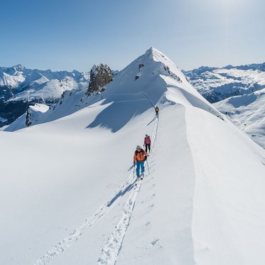 Drei Personen unternehmen eine Skitour auf einem schmalen, verschneiten Berggrat; sie folgen einer Spur im Tiefschnee, umgeben von einem weiten Panorama schneebedeckter Gipfel unter strahlend blauem Himmel.