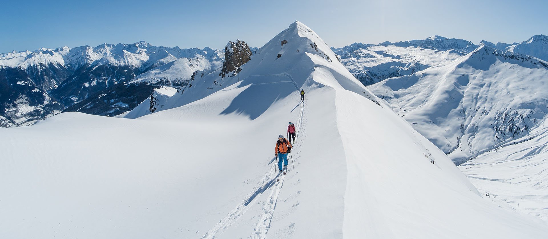 Drei Personen unternehmen eine Skitour auf einem schmalen, verschneiten Berggrat; sie folgen einer Spur im Tiefschnee, umgeben von einem weiten Panorama schneebedeckter Gipfel unter strahlend blauem Himmel.