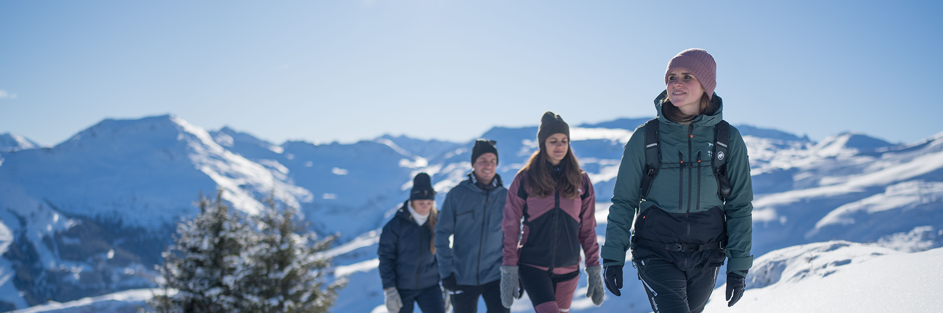 Mehrere Personen unternehmen eine Winterwanderung in Gastein auf einem verschneiten Bergweg; sie tragen warme Kleidung und Rucksäcke, im Hintergrund erstreckt sich ein Panorama schneebedeckter Gipfel unter blauem Himmel.