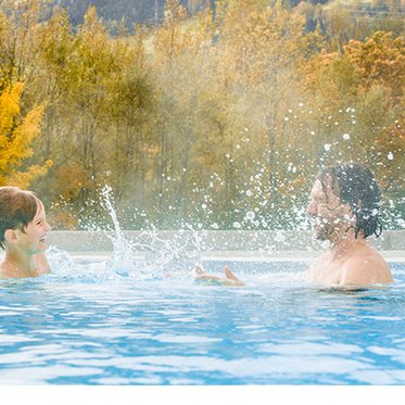 Kind und Vater spritzen sich spielerisch mit Wasser im warmen Außenbecken der Alpentherme Gastein.