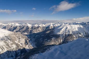 Weites Panorama über das verschneite Gasteinertal mit umliegenden schneebedeckten Berggipfeln unter blauem Himmel.