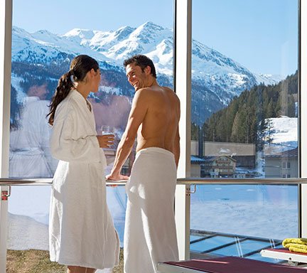 Ein Pärchen steht an dem großen Panoramafenster der Felsentherme in Gastein. Vor Ihnen das Außenbecken, Blick auf Schneebedeckte Gipfel.