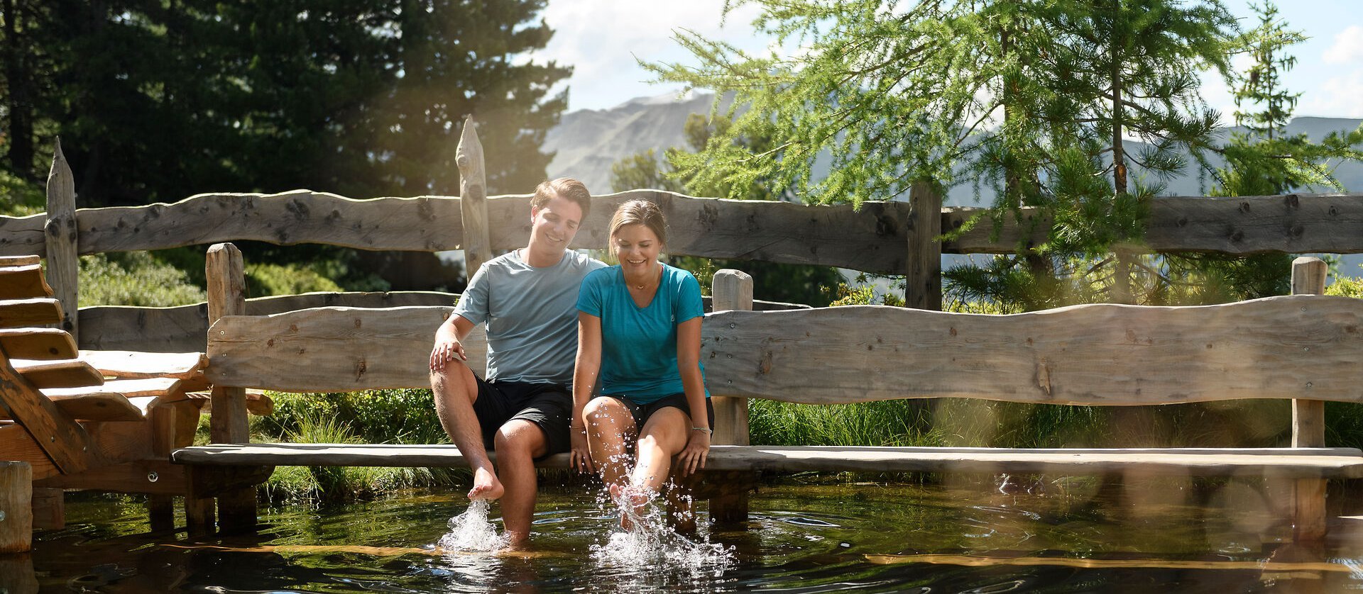 Ein Paar sitzt am Steg in der Zirbenoase und hält die Füße ins Wasser; umgeben von Holzstegen, Bäumen und ruhiger Natur genießen sie eine entspannte Auszeit.