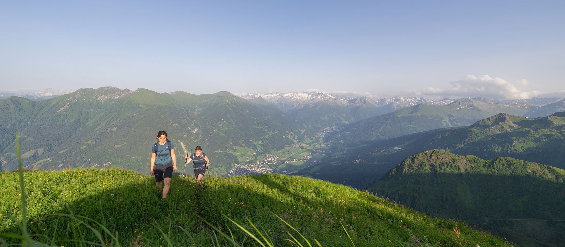 Zwei Personen wandern über einen grünen Bergkamm mit blühender Wiese; im Hintergrund erstreckt sich eine weite Berglandschaft unter klarem Himmel.