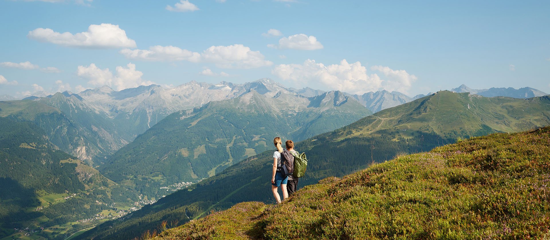 Gastein Trail: Eine Person steht auf einem blühenden Bergkamm und blickt über das weite Gasteinertal mit umliegenden Gipfeln im Salzburger Land unter blauem Himmel mit vereinzelten Wolken.