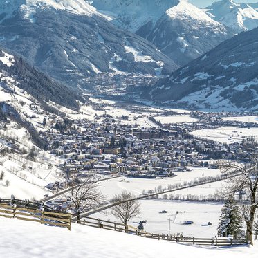 Luftaufnahme von Bad Hofgastein im Winter: Der verschneite Ort liegt im Tal, mittig ragt der Kirchturm empor, umgeben von dicht bebauten Häusern und schneebedeckten Hängen.