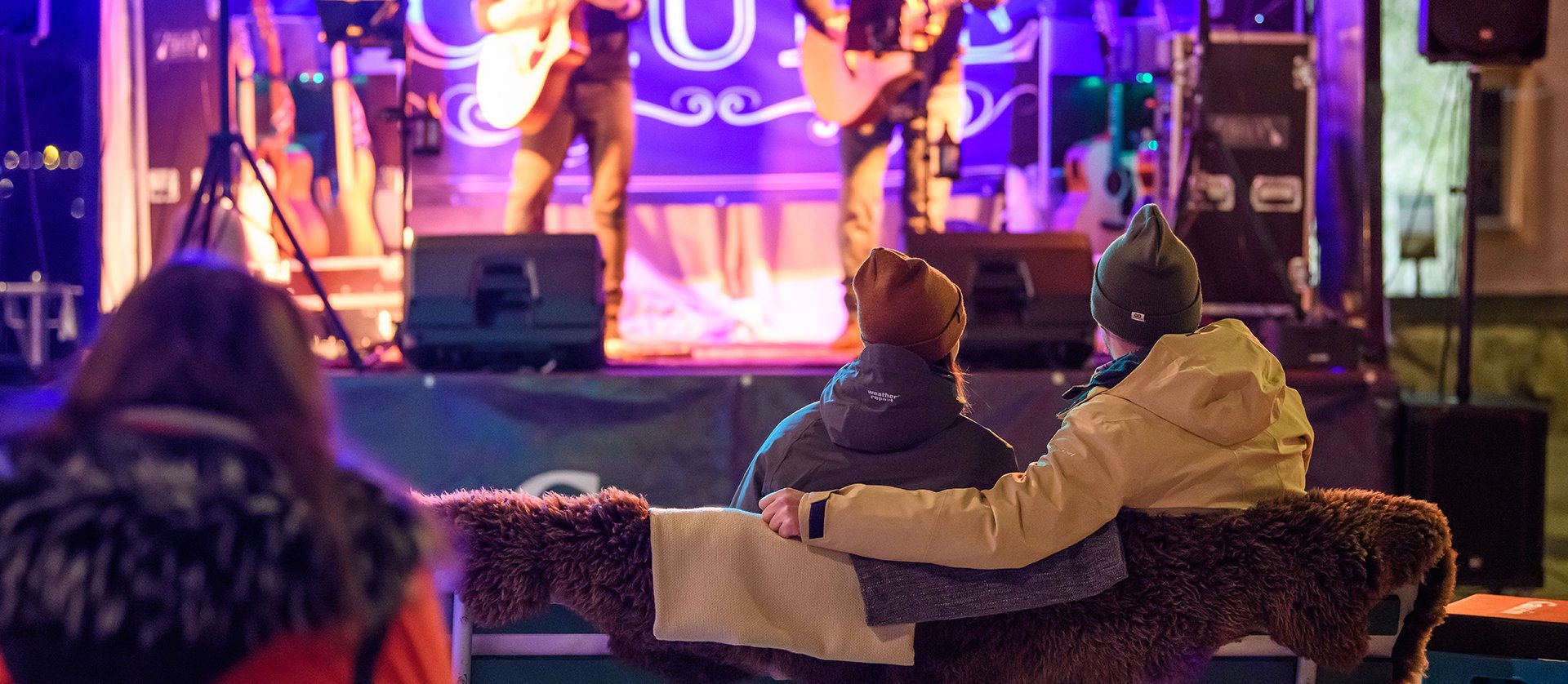 Kleine Konzertbühne in gemütlicher Wohnzimmer-Atmosphäre: Eine Band spielt unter warmem Bühnenlicht, während das Publikum entspannt auf Sofas und Sesseln sitzt und zuhört.