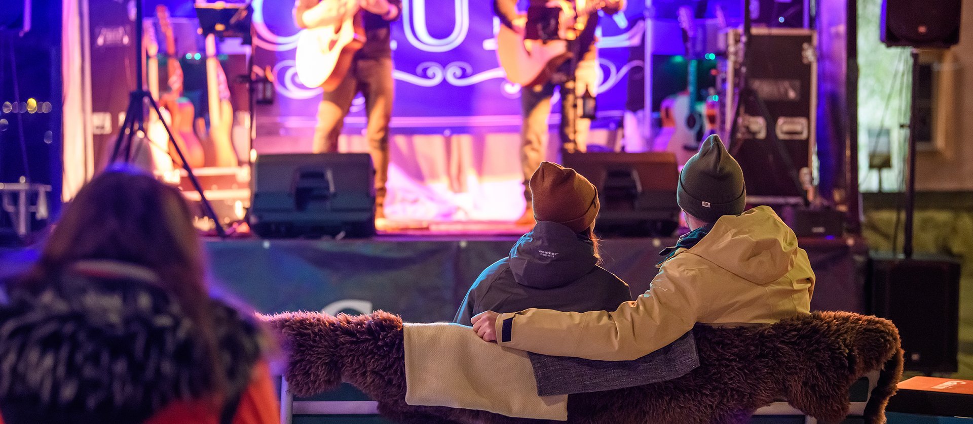 Kleine Konzertbühne in gemütlicher Wohnzimmer-Atmosphäre: Eine Band spielt unter warmem Bühnenlicht, während das Publikum entspannt auf Sofas und Sesseln sitzt und zuhört.