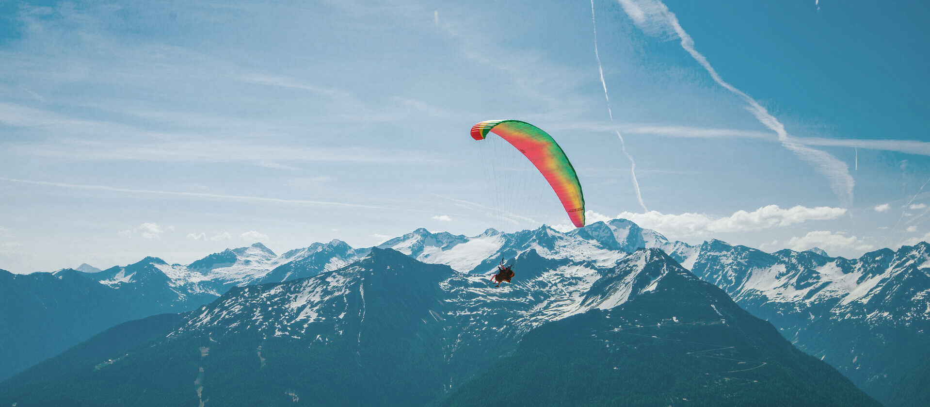 Ein Paragleiter mit buntem Schirm schwebt über einem bewaldeten Berg mit schneebedecktem Gipfel; im Hintergrund erstreckt sich eine weite Berglandschaft unter blauem Himmel mit dünnen Wolkenstreifen.