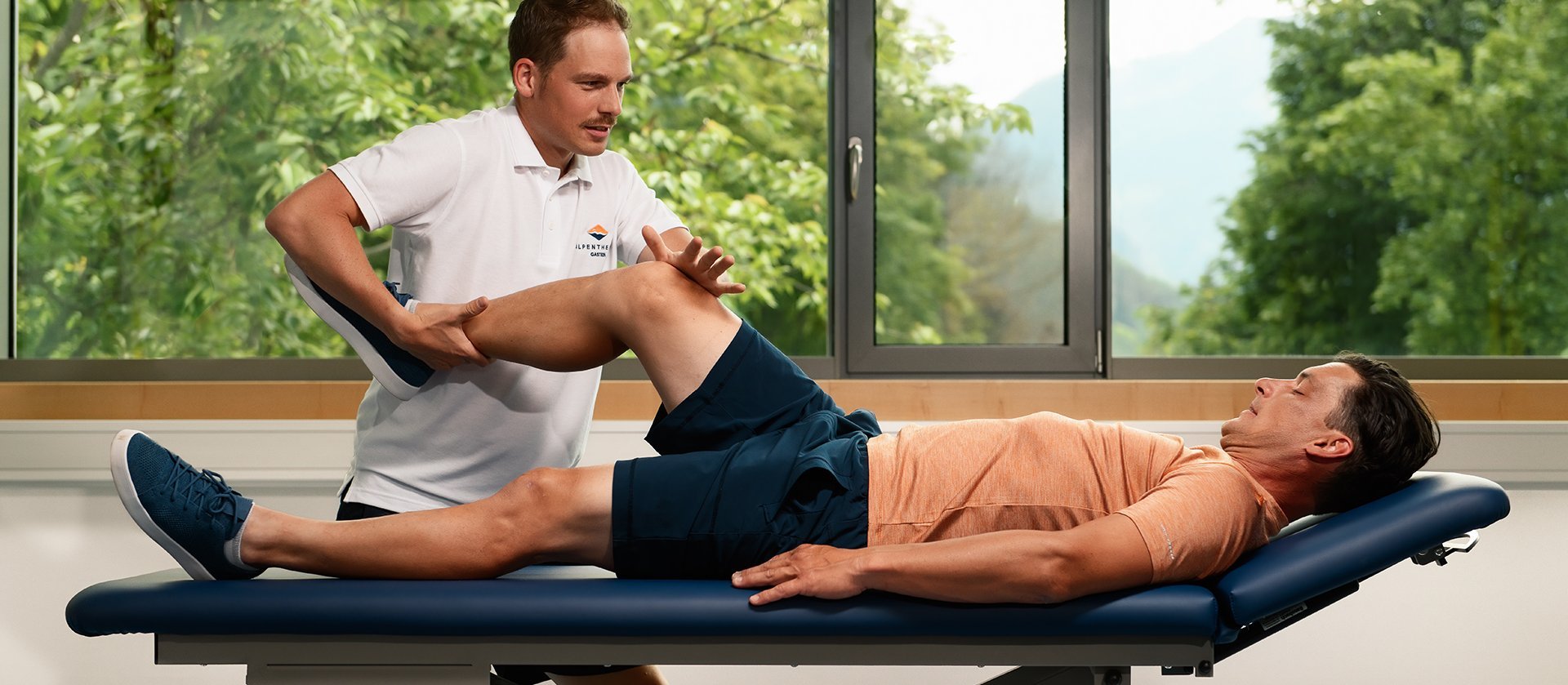 Ein Therapeut führt eine Behandlung an einer liegenden Person auf einer Massageliege durch; die Person liegt entspannt auf dem Rücken, während der Therapeut mit den Händen am Oberkörper arbeitet, im Hintergrund sind große Fenster mit Blick ins Grüne zu sehen.