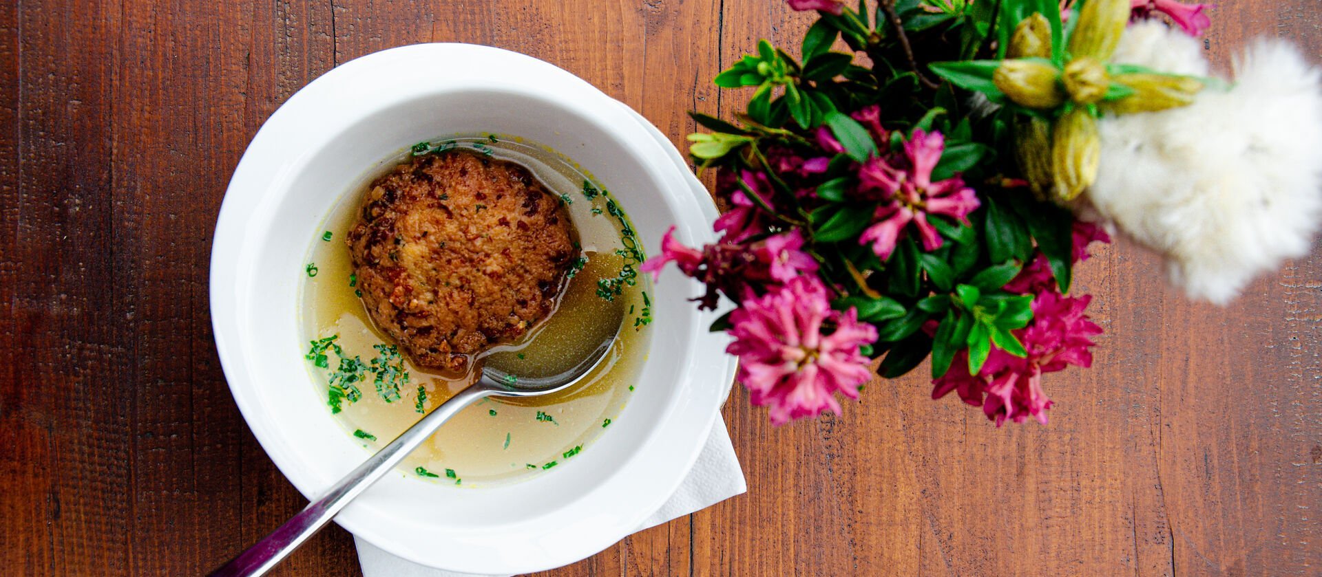 Kaspressknödelsuppe in einer weißen Schüssel auf einem rustikalen Holztisch neben einem bunten Blumenstrauß.