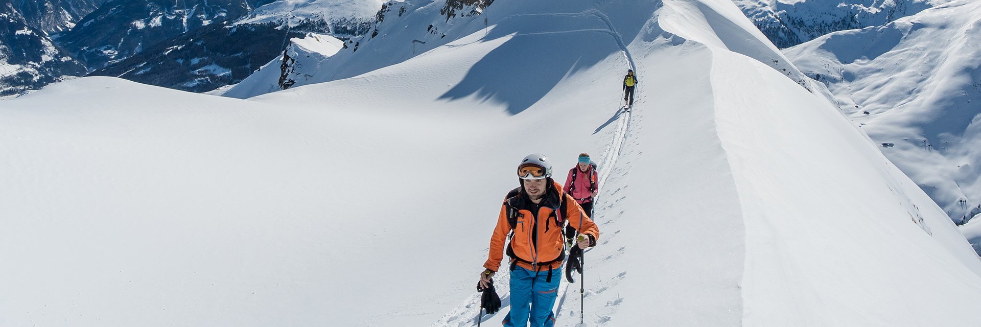 Drei Personen unternehmen eine Skitour auf einem schmalen, verschneiten Berggrat; sie folgen einer Spur im Tiefschnee, umgeben von einem weiten Panorama schneebedeckter Gipfel unter strahlend blauem Himmel.
