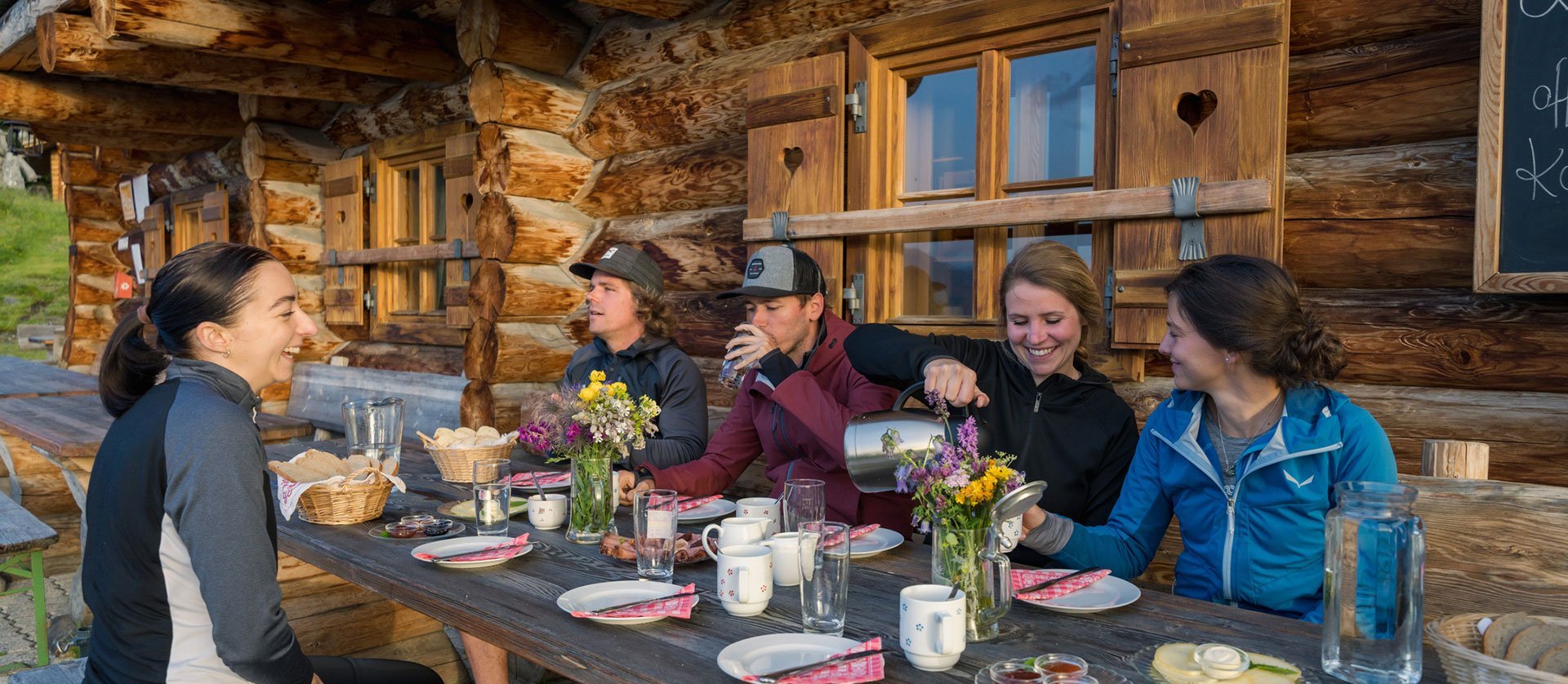 Gruppe von Wanderern sitzt vor einer traditionellen Berghütte und genießt eine gemeinsame Jause mit Brot, Getränken und Blumen auf einem Holztisch.