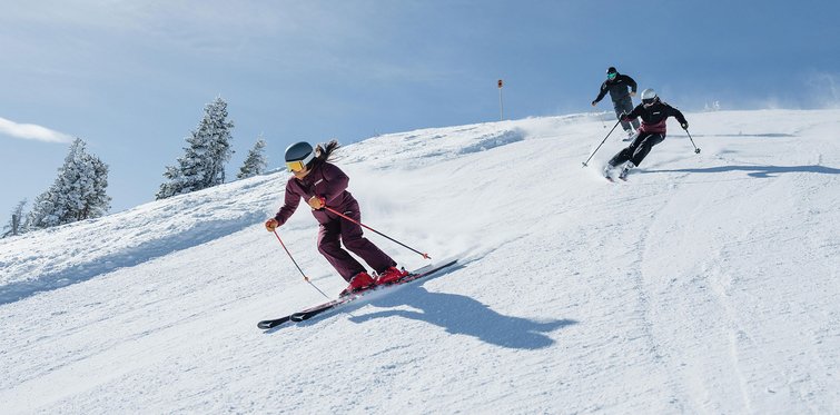Zwei Skifahrer fahren bei sonnigem Wetter eine präparierte Piste hinunter und ziehen Schwünge im Schnee; im Hintergrund sind verschneite Hänge und vereinzelte Bäume zu sehen.