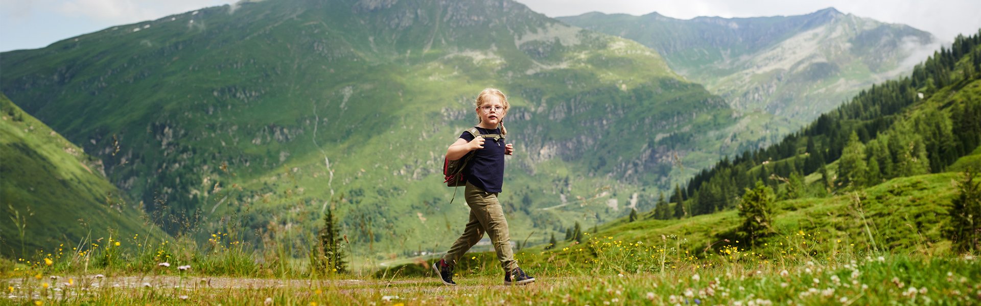 Kleines Kind mit Rucksack wandert über eine blühende Bergwiese vor einer beeindruckenden alpinen Berglandschaft mit grünen Hängen und Gipfeln.
