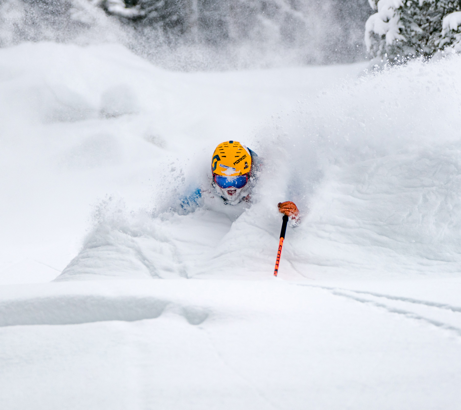 Eine Person mit gelbem Helm fährt durch tiefen Pulverschnee und wirbelt dabei eine große Schneewolke auf; die Szene zeigt eine actionreiche Abfahrt im freien Gelände.